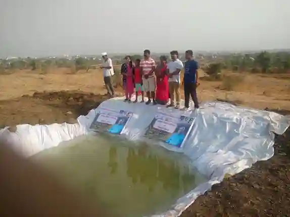 The image shows a group of people standing on the edge of a small water reservoir or pond, which is covered with a white tarp. The setting appears to be a dry, rural area with sparse vegetation in the background. There are several individuals, including men and women, posing together, and some of them are holding signs or banners.The atmosphere seems to be celebratory, possibly indicating the inauguration or completion of a water conservation project.